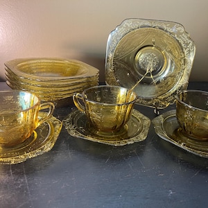 May include: A collection of amber-colored glass teacups, saucers, and plates. The set includes teacups with handles, matching saucers, and square plates. The glass has a decorative embossed floral pattern. A vintage tea set.