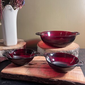 May include: Three red glass bowls with handles. The bowls are on a wooden surface. The largest bowl is in the background and the two smaller bowls are in the foreground.