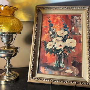 May include: An antique oil lamp with a yellow glass shade and a brass base stands next to a framed floral painting. The painting features white flowers in a vase against a red background, with a gold ornate frame.