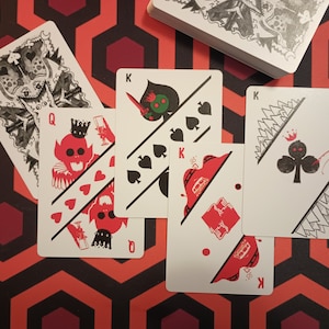 May include: A deck of playing cards with unique illustrations. Several cards are fanned out, showcasing red and black designs on a white background. The cards feature a skull-like Queen of Hearts and a King of Spades with a sword.