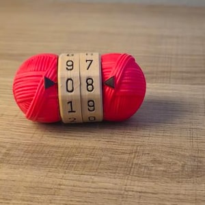 以下が含まれることがあります： 数字の9、7、0、8、1、9が表示されたコンビネーションロックが付いた赤い毛糸玉。ロックは明るい色の木材でできています。毛糸玉は木製の表面に置かれています。