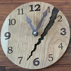 May include: A round wooden clock with a light brown face and dark brown numbers and hands. A dark brown jagged line runs vertically across the face. The clock hands point to approximately 11:58. Home decor.