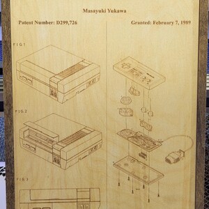 Puede incluir: Obra de arte enmarcada en madera con un diagrama grabado con láser del sistema Nintendo Entertainment. La obra de arte incluye la información de la patente del sistema, diagramas explosionados y el texto "Nintendo Entertainment System".