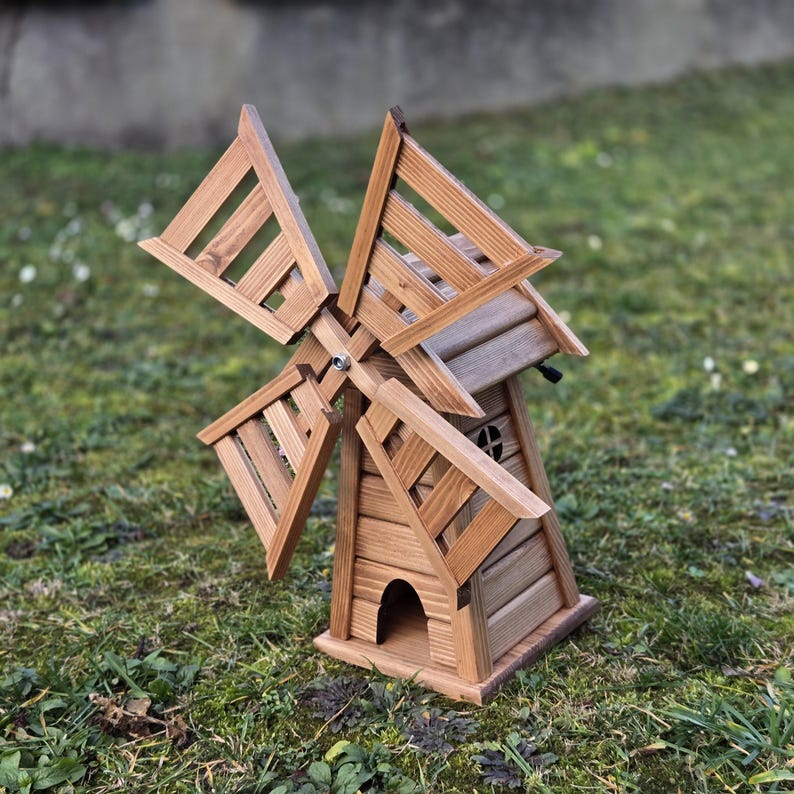 May include: A wooden windmill garden ornament featuring a natural brown finish, four slatted rotating blades attached to a central hub, and a small arched opening at the base of the tower structure, showcasing a detailed horizontal plank design.