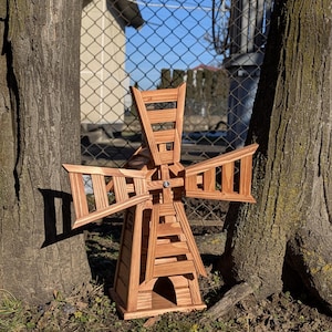 May include: A wooden windmill garden ornament featuring a natural brown finish with visible wood grain. The structure has a tapered base with an arched opening and four rotating blades attached to a central hub with a visible metal bolt.