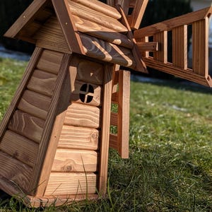 May include: A wooden windmill model constructed from light brown horizontal logs with a peaked roof and a small circular window. The structure features a large, rotating-style blade assembly attached to the side, crafted from matching wood slats.