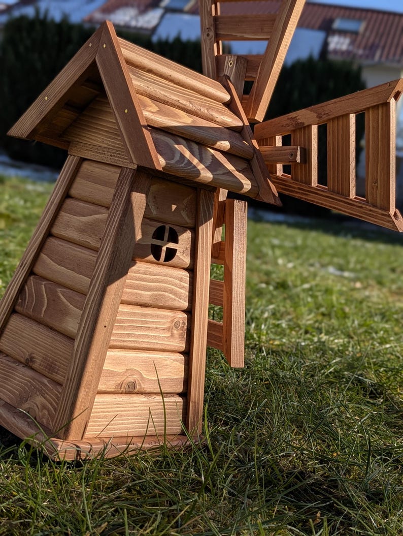 May include: A wooden windmill model constructed from light brown horizontal logs with a peaked roof and a small circular window. The structure features a large, rotating-style blade assembly attached to the side, crafted from matching wood slats.