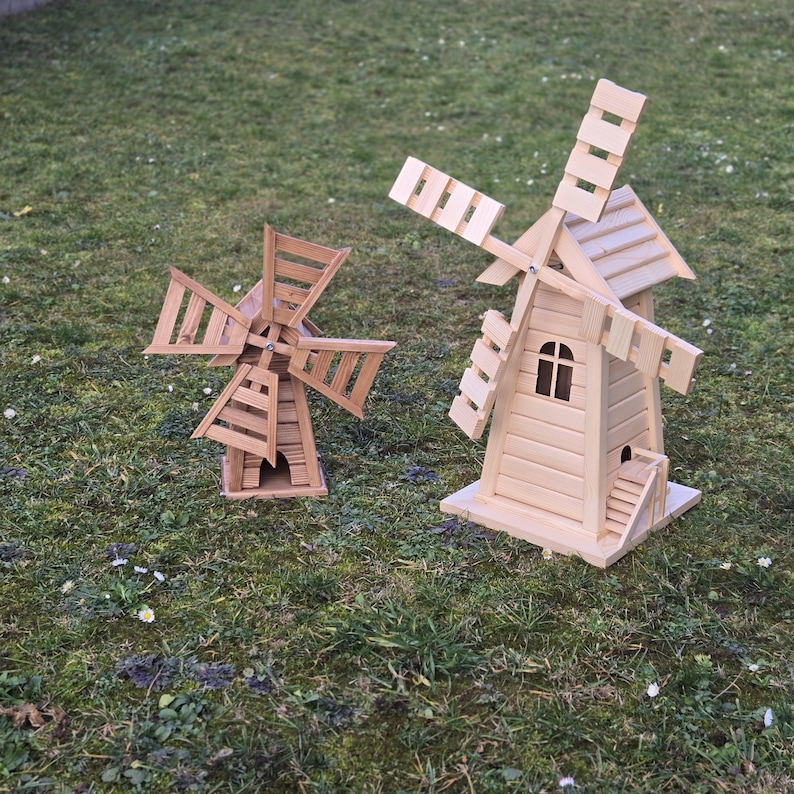 May include: Two light-coloured wooden windmill models with rotating blades. The larger windmill features a peaked roof, a small arched window, and a set of stairs leading to a doorway. The smaller windmill has a simpler structure with four blades.