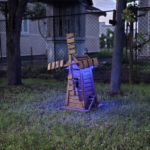 May include: A wooden windmill structure featuring horizontal slats and a rotating blade assembly. The base of the windmill is illuminated by bright blue LED lights that cast a glow onto the surrounding grass, highlighting the wood grain texture.