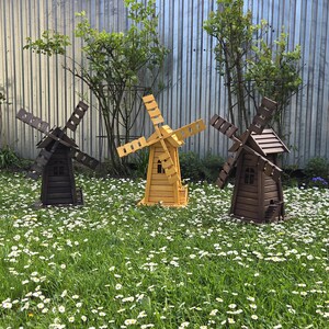 May include: Three wooden windmill garden ornaments standing in a field of grass and white daisies. The left and right windmills are dark brown, while the centre windmill is a light natural wood colour. Each features a roof, blades, and a small door.