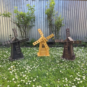 May include: Three wooden windmill garden ornaments standing in a grassy field. Two windmills are dark brown with four blades and a small window, while the central windmill is a lighter yellow-orange wood with a matching four-blade design.