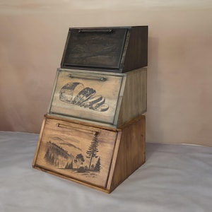 Op de afbeelding: Drie houten brooddozen op elkaar gestapeld. De bovenste doos is zwart, de middelste doos is lichtbruin met een zwart-witte illustratie van brood en de onderste doos is donkerbruin met een zwart-witte illustratie van een bos scène.