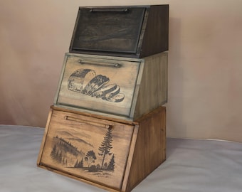 Könnte beinhalten: Drei übereinander gestapelte Holzbrotboxen. Die obere Box ist schwarz, die mittlere Box ist hellbraun mit einer Schwarzweißillustration von Brot und die untere Box ist dunkelbraun mit einer Schwarzweißillustration einer Waldszene.