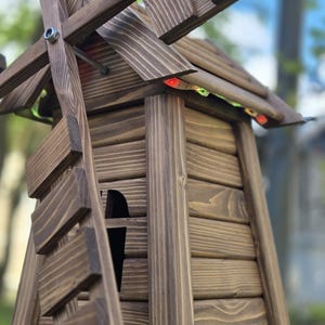May include: A dark brown wooden windmill structure featuring horizontal plank siding, a tapered body, and rotating blades attached with a silver metal bolt. Small red and green LED light strips are tucked under the roof eaves of the structure.