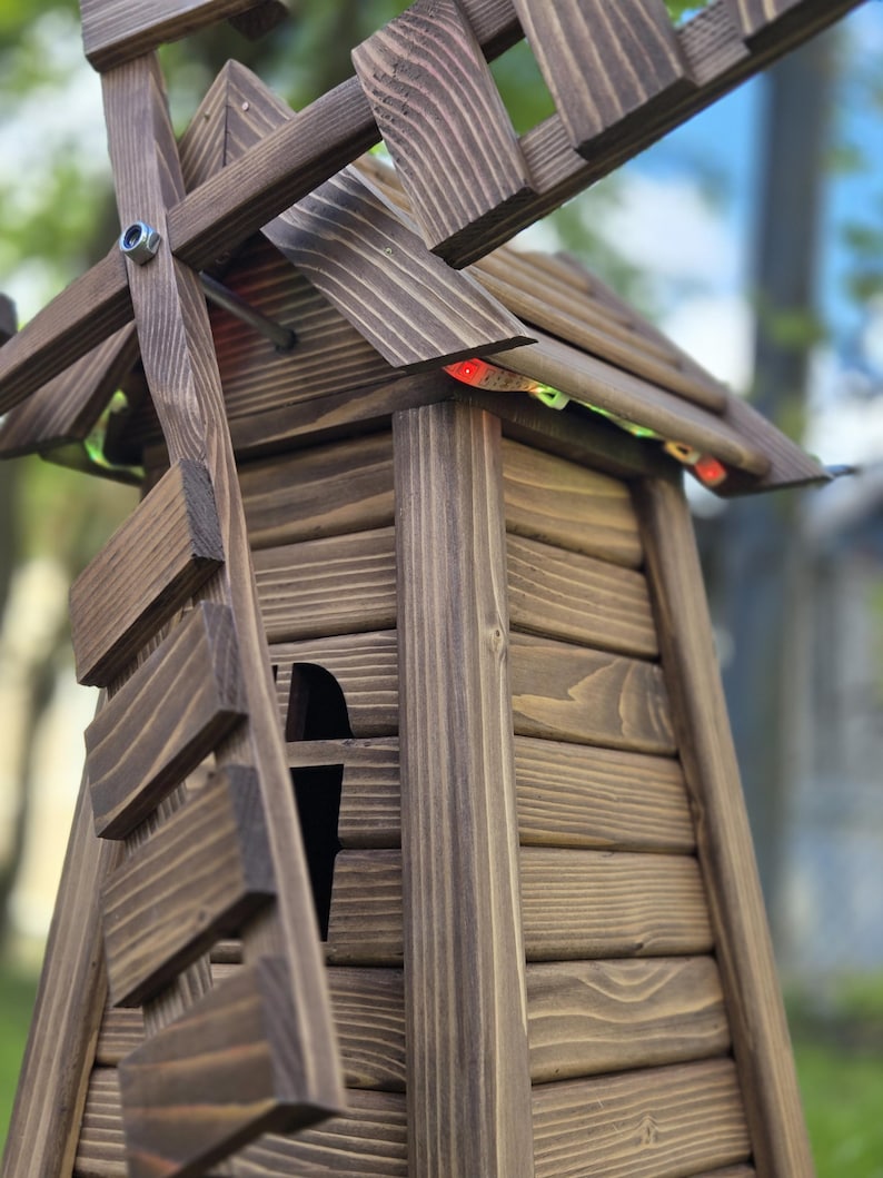 May include: A dark brown wooden windmill structure featuring horizontal plank siding, a tapered body, and rotating blades attached with a silver metal bolt. Small red and green LED light strips are tucked under the roof eaves of the structure.