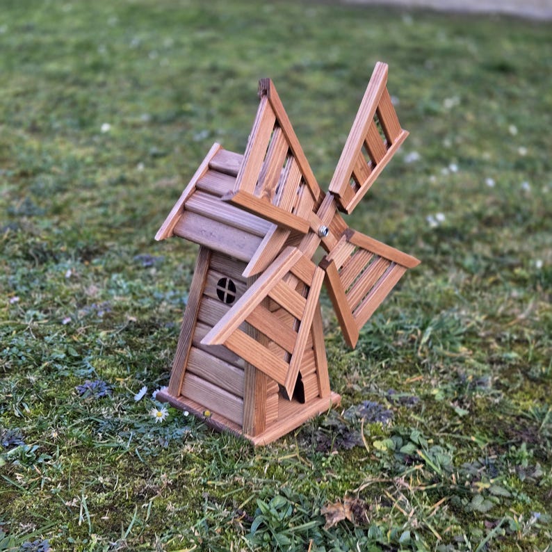 May include: A brown wooden windmill model featuring a slatted main structure with a small circular window and four rotating blades attached to the front. The windmill is constructed from horizontal planks and has a pointed roof design.