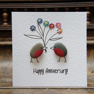 May include: A handmade greeting card featuring a painted design of two birds holding a bouquet of flowers. The card reads "Happy Anniversary!"