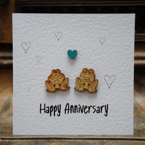 May include: A white card with the words "Happy Anniversary" written in black ink. Two wooden frog figurines are attached to the card, one with a bow tie. There are several small hearts drawn in pencil around the frogs.