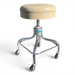 May include: A round stool with a beige padded seat and a chrome metal base. The stool features an adjustable height mechanism with a central screw, three curved legs, and black caster wheels. A small blue rectangular label is on the central hub.