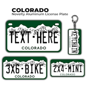 May include: A collection of Colorado-themed novelty aluminum license plates and a keychain. The plates are white with a green border and feature a mountain range design. Text on the plates includes "TEXT-HERE", "3X6-BIKE", and "2X4-MINI". The keychain reads "KEYCHAIN".
