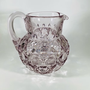 May include: A clear glass pitcher with a diamond pattern and a light purple hue. The pitcher has a handle and a spout.