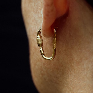 May include: A gold-coloured, carabiner-style earring is worn on the ear. The earring has a hinged design with a screw-like closure. The background is a solid black.