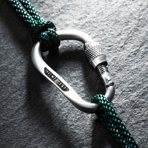 Könnte beinhalten: Nahaufnahme eines silbernen Karabiners mit Schraubverschluss, verbunden mit einem dunkelgrünen Seil. Das Seil ist um den Karabiner geknotet. Der Hintergrund ist eine dunkle, strukturierte Oberfläche. Der Karabiner trägt den Schriftzug "AMI GEAR".
