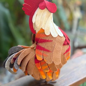 May include: A decorative rooster figurine crafted from layered felt pieces. The bird features a bright red comb and wattle, cream-colored neck feathers, and wings with overlapping brown, orange, and red felt segments. The tail includes gray accents.