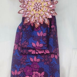 May include: A purple tie with a floral pattern, accented by a large pink and gold brooch. The tie is adorned with pink and blue flowers and leaves. The tie is worn with a white collared shirt.