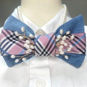 May include: A denim and plaid bow tie with pearl accents. The bow tie features a blue denim base with a pink, blue, and black plaid center. Delicate gold wire and faux pearls adorn the bow tie, adding a touch of elegance. The bow tie is set against a white shirt.