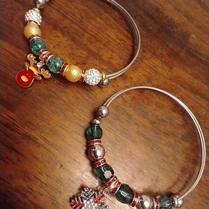 May include: Two silver bangle bracelets with green and red beads, one with a reindeer charm and the other with a snowflake charm.