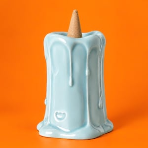 May include: A light blue ceramic candle-shaped incense burner with a glossy finish and sculpted wax drip details. A single tan-colored conical incense cone is positioned upright in the center of the top opening of the ceramic holder.