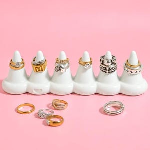 May include: A white ceramic ring holder with six cone-shaped prongs displaying various gold and silver rings, including one with an H emblem and one with a cross motif. Four additional rings are placed on the surface in front of the holder.