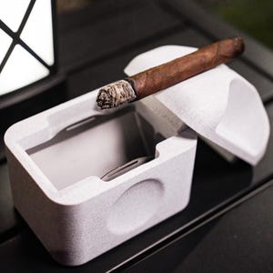 May include: A white ceramic cigar ashtray with a lid. The lid is open and a partially smoked cigar is resting on top of the lid.