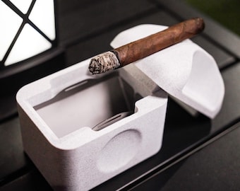 May include: A white ceramic cigar ashtray with a lid. The lid is open and a partially smoked cigar is resting on top of the lid.
