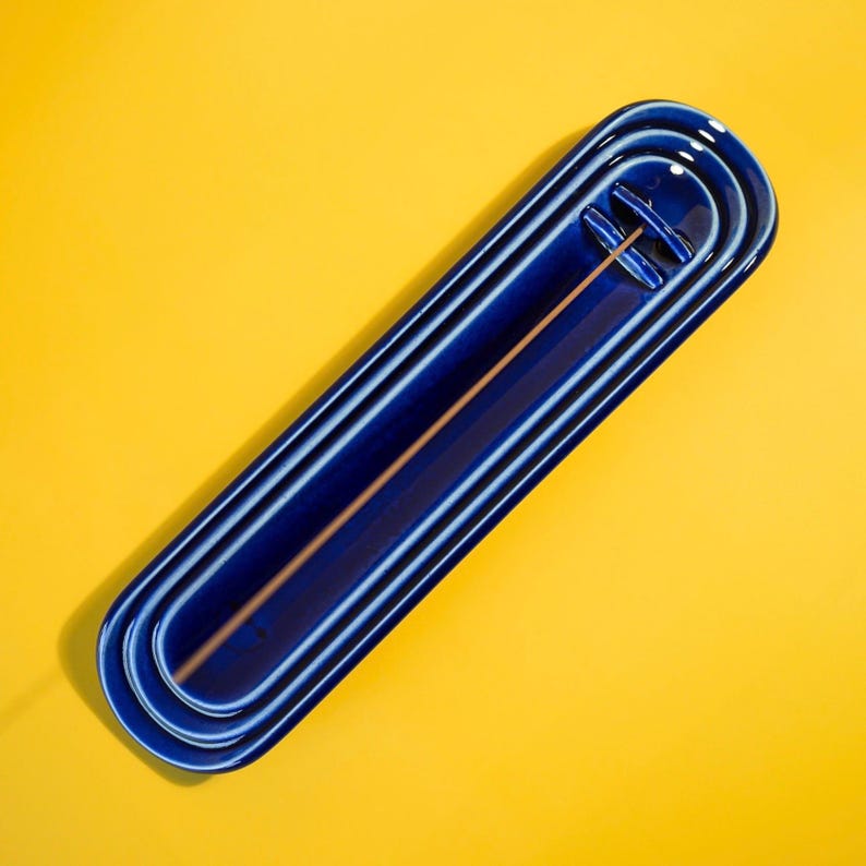 May include: A glossy, deep blue ceramic incense holder with an elongated, rounded rectangular shape. The tray features a central channel and a small, raised bridge-like structure at one end designed to hold a single thin wooden incense stick.
