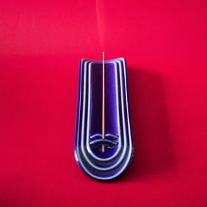 May include: A deep purple ceramic incense holder with a glossy finish and concentric U-shaped ridges. A thin, light brown incense stick is inserted into the small circular opening at the base, with a faint wisp of smoke rising vertically from the tip.