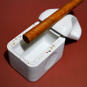 May include: A rectangular white concrete ashtray with a speckled terrazzo finish featuring multicolored stone chips. A brown cigar rests across the top of the ashtray, which includes a curved, elevated notch designed to hold the cigar in place.