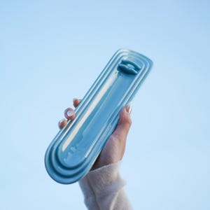 May include: A long, narrow, light blue ceramic tray with a glossy finish and rounded ends. The tray features a series of concentric, raised, oval-shaped ridges that frame a central recessed area, with a small, raised, horizontal bar near one end.