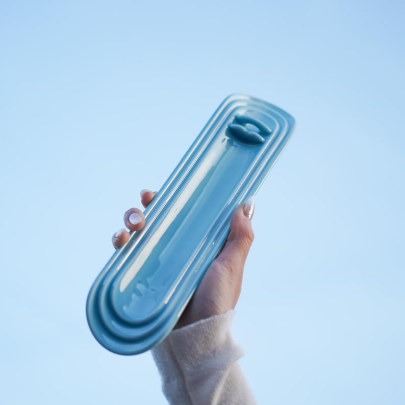 May include: A long, narrow, light blue ceramic tray with a glossy finish and rounded ends. The tray features a series of concentric, raised, oval-shaped ridges that frame a central recessed area, with a small, raised, horizontal bar near one end.