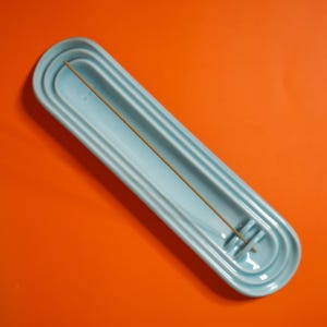May include: A light blue ceramic incense holder with a long, oval shape and concentric grooved detailing. The holder features a small, raised circular notch at one end designed to secure a single, thin, brown wooden incense stick in place.