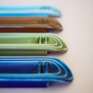 May include: A stack of four elongated ceramic spoon rests in varying colors including light blue, brown, sage green, and deep royal blue. Each piece features a smooth, glossy finish with raised parallel ridges and a curved end design.