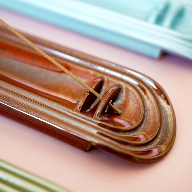 May include: A glossy, elongated, brown ceramic incense holder with a tiered, arched design at one end. A thin, light brown incense stick is inserted into the small, curved holder feature, extending diagonally across the tray's surface.