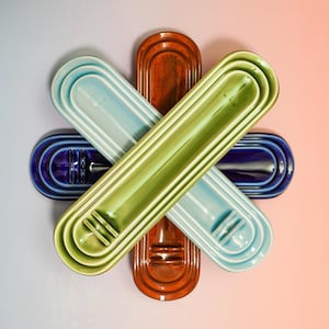 May include: A collection of four elongated, oval-shaped ceramic trays arranged in a star pattern. The trays feature a glossy finish in shades of light blue, deep blue, olive green, and burnt orange, each with a raised, ribbed design at one end.