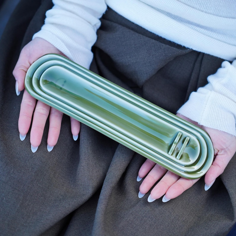 May include: A long, rectangular, olive green ceramic tray with rounded ends and a glossy finish. The tray features a series of concentric, raised ridges that follow the outer shape, with two small, parallel vertical bars near one end.