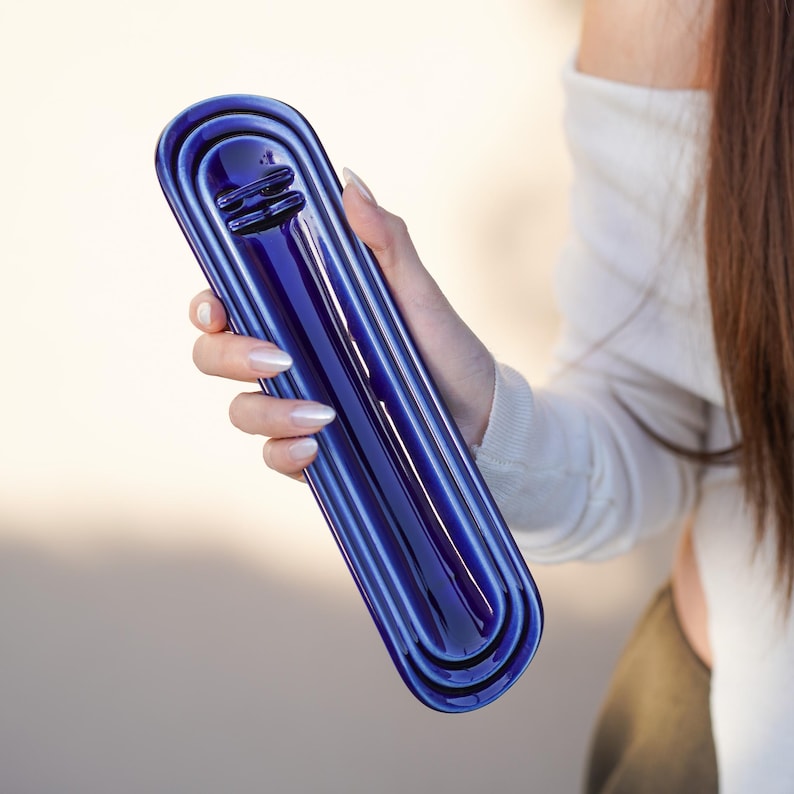 May include: A long, narrow, deep blue ceramic tray with a glossy finish and rounded ends. The tray features a series of concentric, recessed oval channels that run the length of the object, creating a layered, stepped appearance in the center.