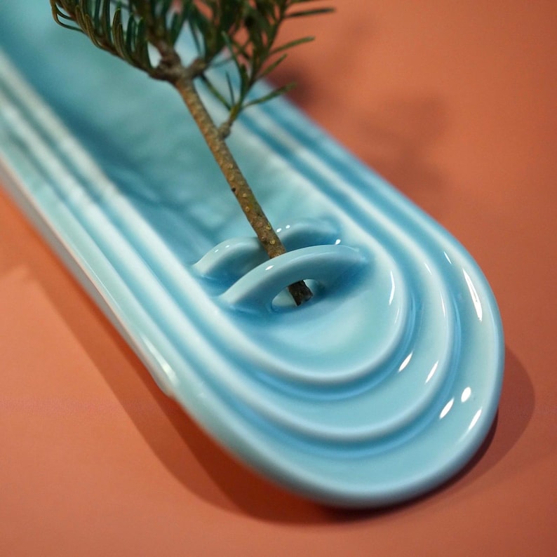 May include: A light blue ceramic tray with a long, oval shape and concentric, raised ridges. A small, arched bridge feature is integrated into the surface, holding a single, thin, brown-stemmed evergreen branch with green needles.