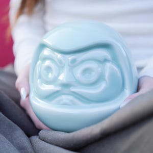 May include: A light blue Daruma doll with a detailed face. The ceramic doll has a smooth, glossy finish and is held in place by a person. The Daruma doll is a traditional Japanese good luck charm, often used for goal setting and motivation.