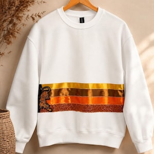 May include: A white long-sleeve sweatshirt featuring a horizontal band across the chest with three distinct stripes in yellow, brown, and orange, accented by a glittery copper texture and a black tiger graphic on the left side.