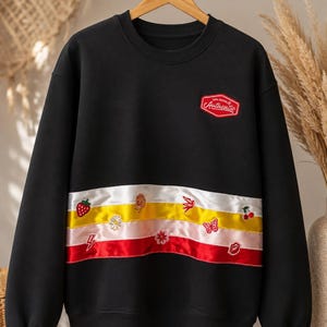 May include: A black long-sleeve sweatshirt featuring a red patch on the chest with the word Authentic in white script. A horizontal band across the midsection displays three satin stripes in white, yellow, and red, decorated with small embroidered icons.
